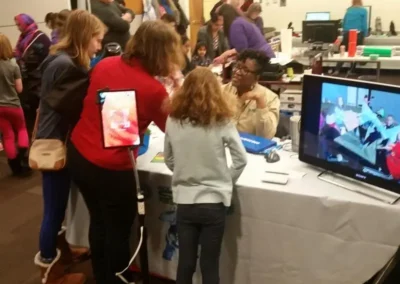 Children and parents gather at a STEM event table where a volunteer demonstrates technology on a tablet and a large screen.